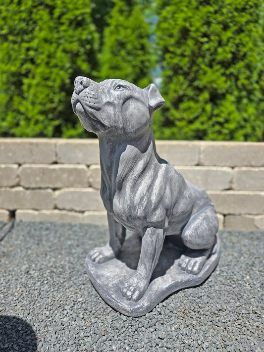 A gray stone-style dog statue, shaped like a muscular pit bull sitting and looking upward, placed outdoors on a gravel surface with a stone wall and green hedges in the background.