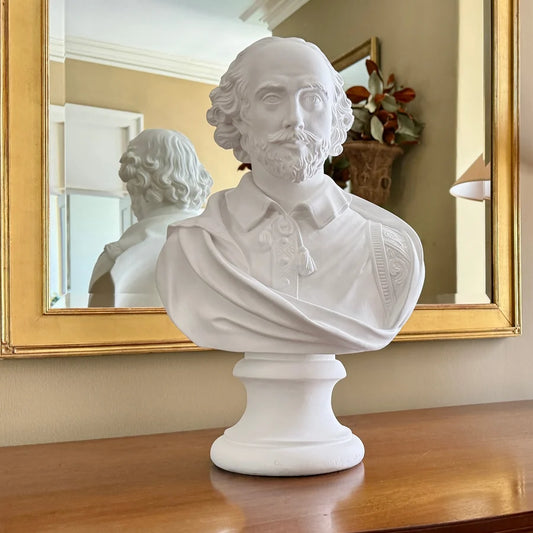 A white classical-style bust sculpture of William Shakespeare on a pedestal, displayed on a wooden surface in front of a gold-framed mirror.