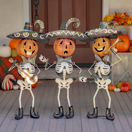 Three Halloween skeleton yard decorations with jack-o'-lantern heads and large sombreros, posed on a gray wooden porch in front of a brown door with pumpkins and fall decor in the background.