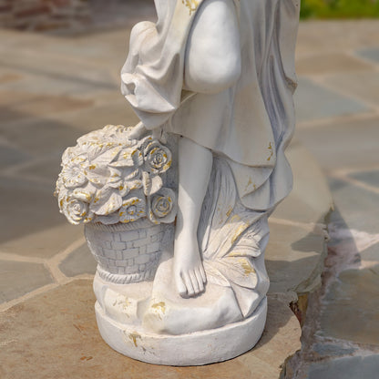 A close-up of a white garden statue depicting a draped female figure with a basket of roses, with subtle gold accents, set on a stone patio.
