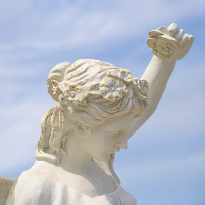 A white stone or plaster sculpture of a classical female figure with one arm raised, holding a flower, set against a soft blue sky with light clouds.