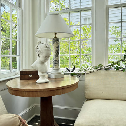Round wooden table with decorative items in a sunlit room with large windows.