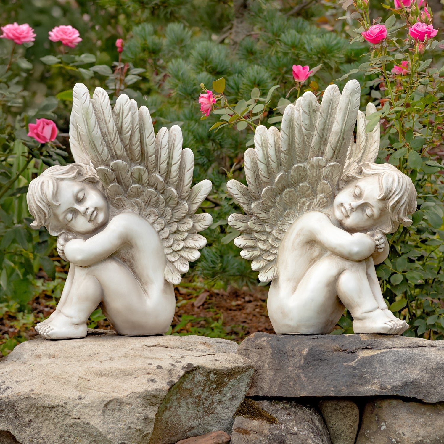 Two white angel garden statues with large feathered wings, shown seated on rocks in a garden with green foliage and pink flowers in the background.