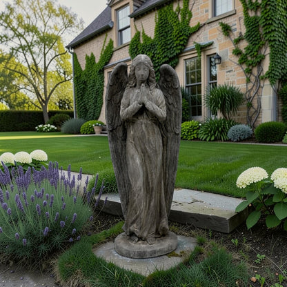 A weathered gray stone angel garden statue with outstretched wings and clasped hands, standing on a circular base in a landscaped lawn with lavender, hydrangeas, and a stone house with ivy in the background.