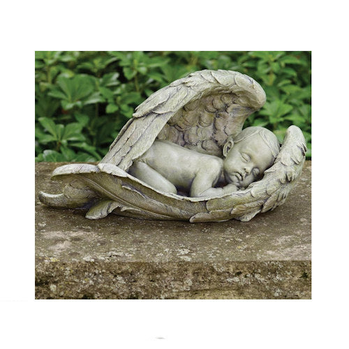 A stone angel garden statue with a sleeping child nestled inside folded wings, displayed outdoors on a stone surface with green foliage in the background.