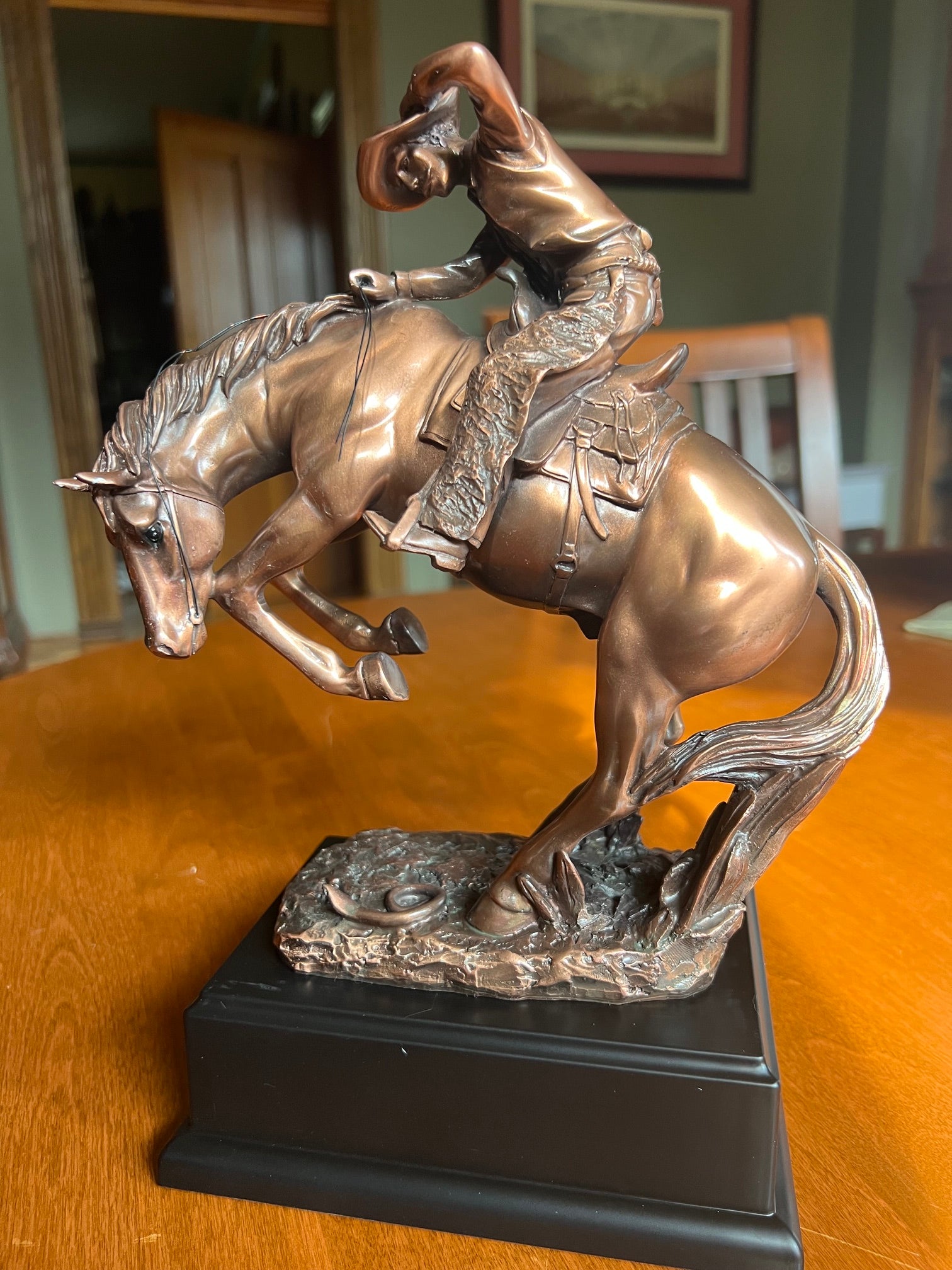 Bronze-colored metal sculpture of a cowboy riding a bucking horse, mounted on a black rectangular base, displayed on a wooden table in an indoor setting.