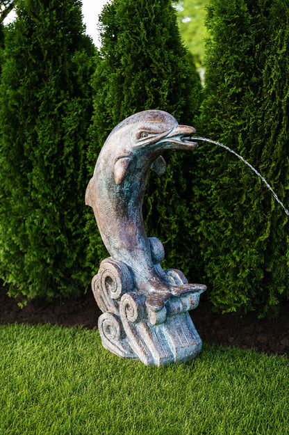 A decorative dolphin fountain sculpture with a weathered bronze and blue-gray finish, shown outdoors on green grass with tall dark green hedges in the background. Water is visible spraying from the dolphin's mouth.