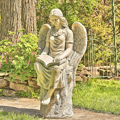 Statue of an angel reading a book in a garden setting with trees and flowers.