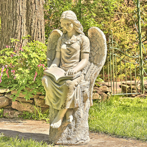 Statue of an angel reading a book in a garden setting with trees and flowers.