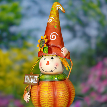 A decorative autumn scarecrow garden ornament with a pumpkin-shaped body, orange hat, sunflower accent, and a small 'WELCOME' sign, photographed against a blurred outdoor garden background.