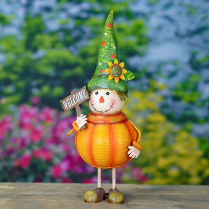 A decorative scarecrow garden statue shaped like a pumpkin, wearing a pointed green hat with a sunflower accent and holding a small 'WELCOME' sign, standing on a wooden surface with a blurred garden background.