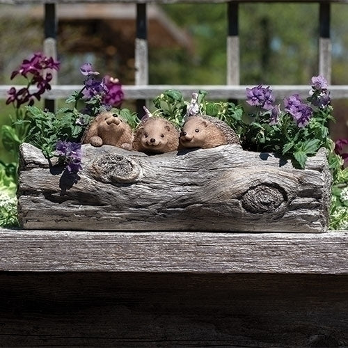 Three small hedgehog figurines sitting on a wooden log with purple flowers in a garden setting.