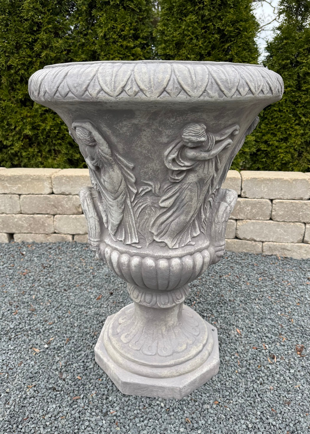 A large gray stone garden urn with classical relief carvings of draped figures, set outdoors on a gravel surface with a stone wall and green trees in the background.