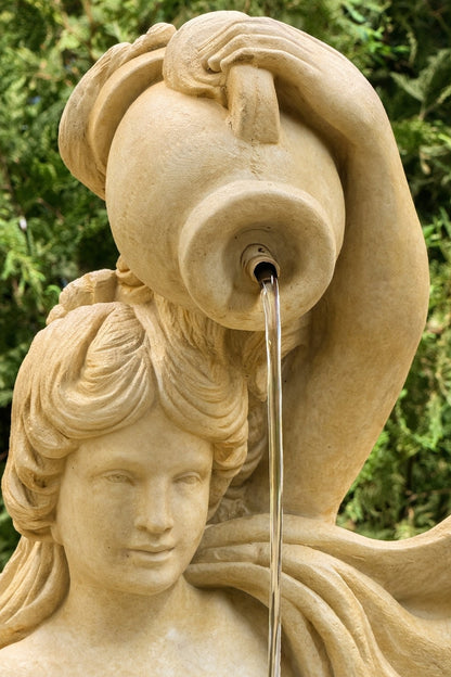 Stone sculpture of a woman with flowing hair and a vase, with water spouting from it, against a green background.