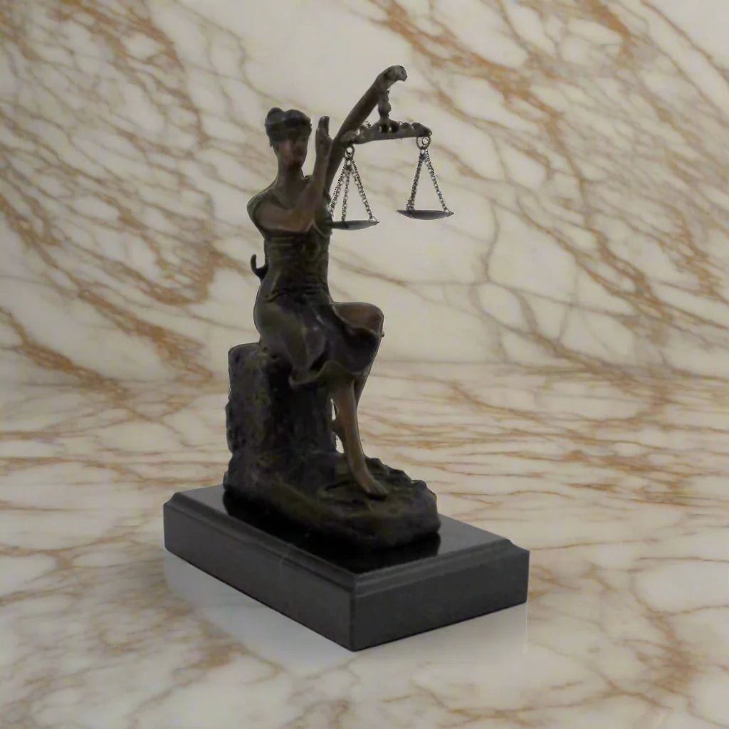 Blind lady justice, sitting down, with her hand waving above her scales of justice.  On a black marble base, with a white veined marble background.