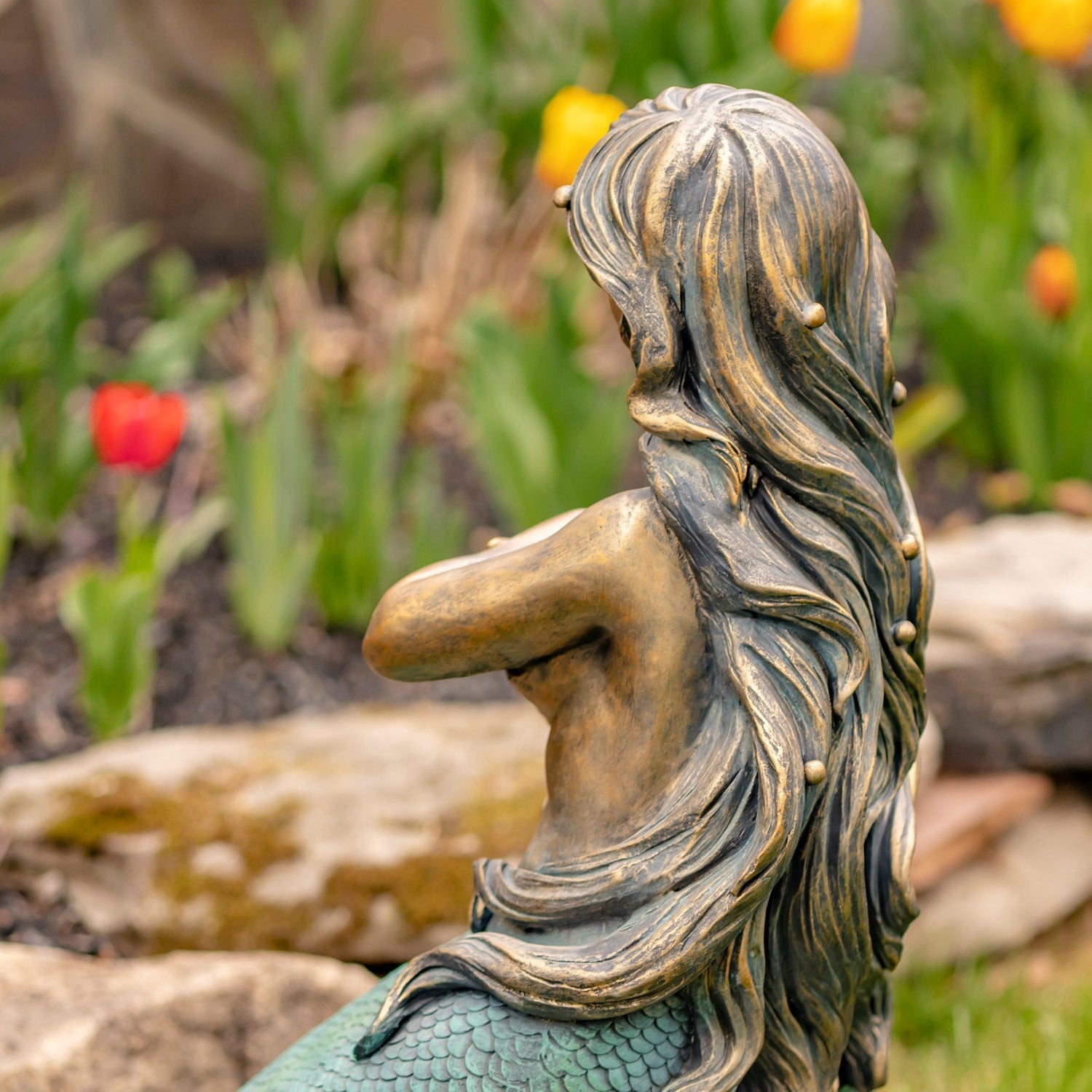 A bronze-finished mermaid garden statue shown from the back, featuring long flowing hair with small bead details and a green scaled tail, set outdoors in a garden with blurred flowers and rocks.