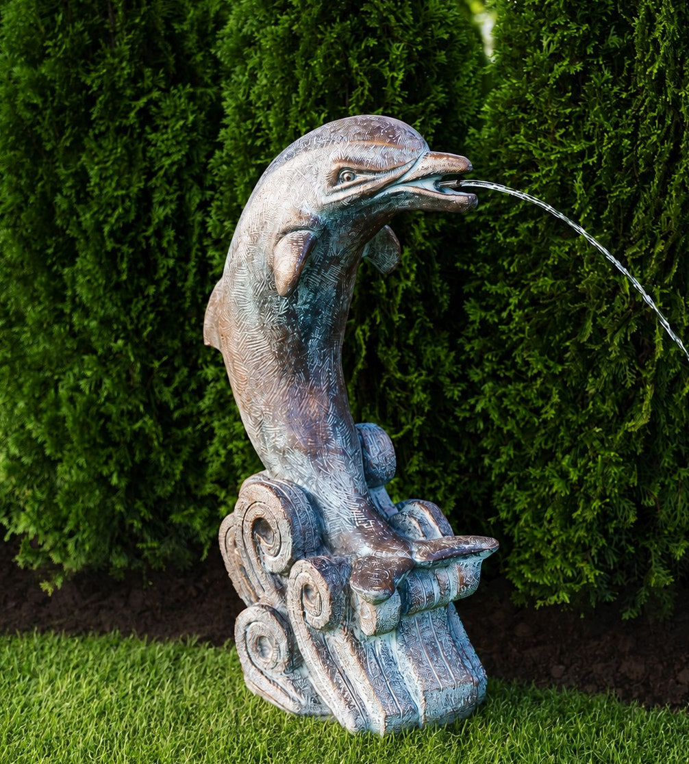 A sculpted dolphin fountain statue with a weathered bronze and blue-green patina finish, perched on a wave-like base and spraying water, set outdoors against a backdrop of green hedges and grass.