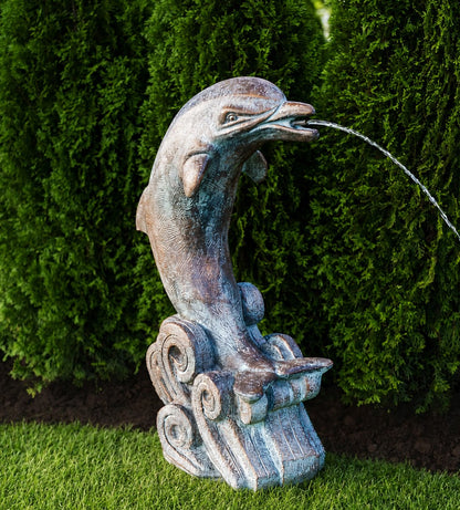 A sculpted dolphin fountain statue with a weathered bronze and blue-green patina finish, perched on a wave-like base and spraying water, set outdoors against a backdrop of green hedges and grass.