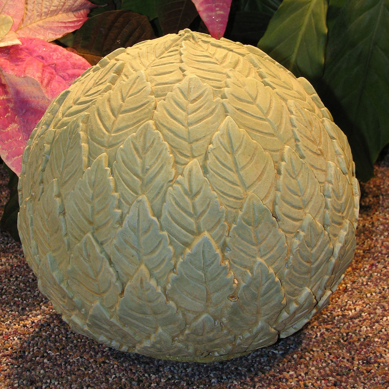 A decorative spherical garden sculpture with a textured leaf pattern, shown outdoors on gravel with plants in the background.