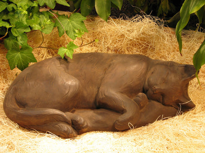 A realistic brown cat sculpture curled up sleeping on a bed of straw, framed by green ivy leaves.